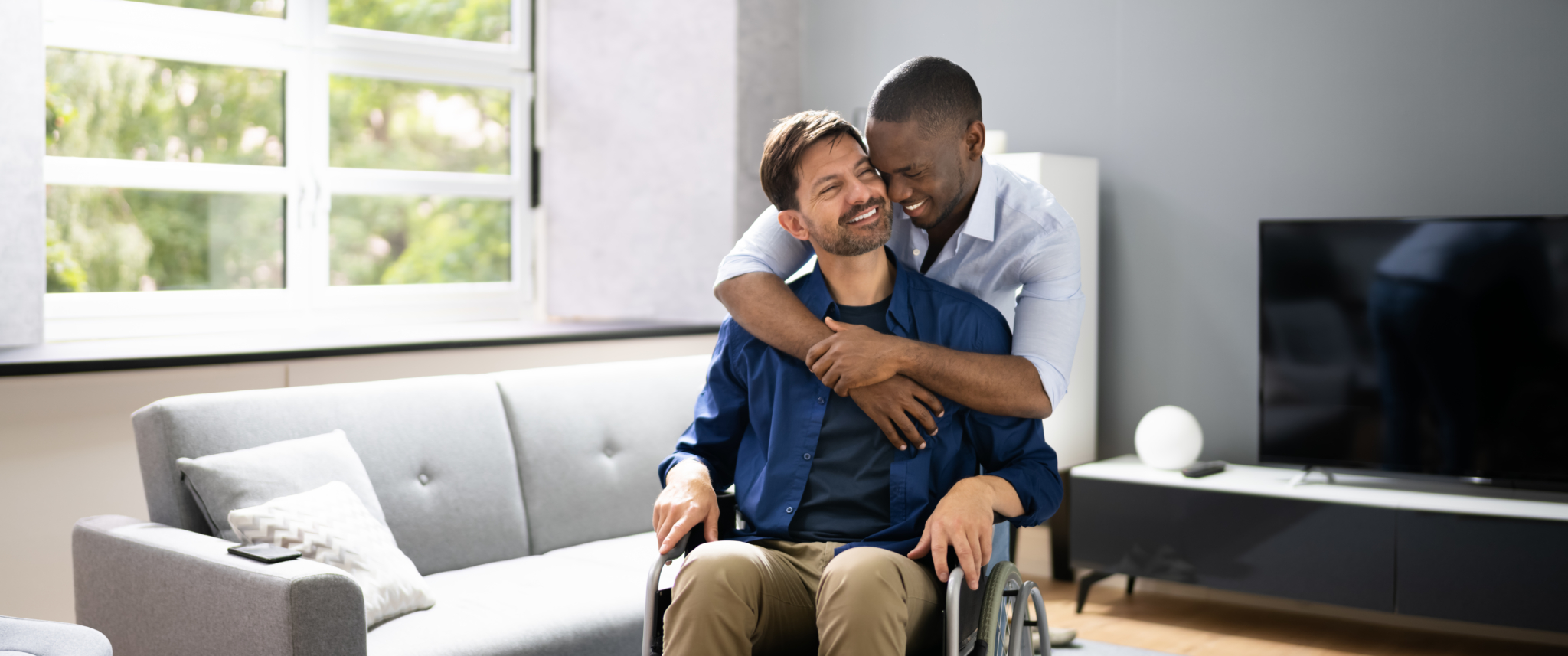 Loving gay couple embrace. One man sits in a wheel chair while his partner gives him a hug. Both are smiling