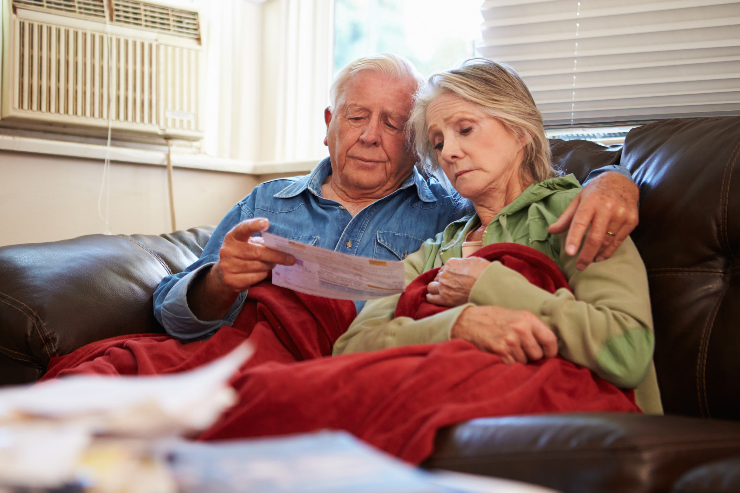 Worried Senior Couple Sitting On Sofa Looking At Medicaid notice Under A Blanket
